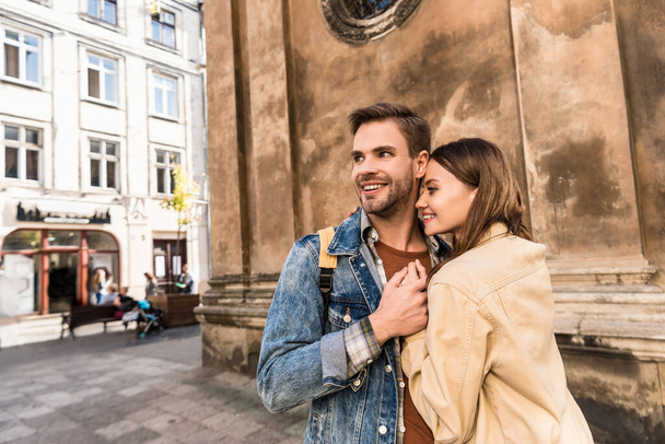 Selective focus of girlfriend and boyfriend looking away, holding hands and smiling near wall in city - Zdjęcie, obraz