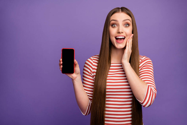 Omg look new device. Astonished surprised girl hold cellphone present incredible ad promotion impressed scream wow omg wear sweater isolated over purple color background - Fotografie, Obrázek