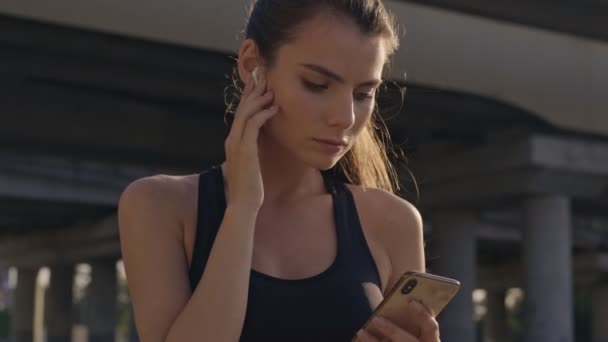A slim sporty young woman is using her earphones while holding a smartphone outdoors near the bridge in the city - Video