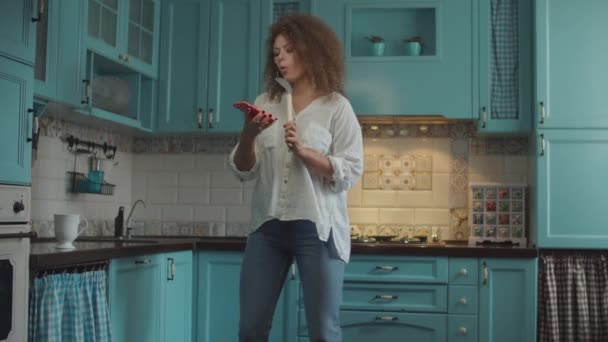 Smiling curly 20s woman in jeans dancing happily with scoop as microphone and mobile phone in hands on home blue kitchen.  - Imágenes, Vídeo