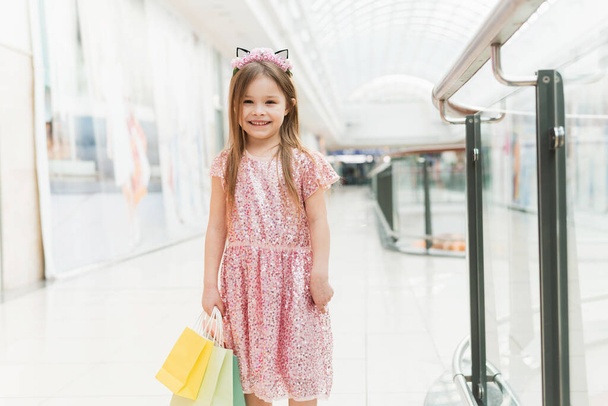 Pequeña chica de moda con paquetes en un gran centro comercial. Una niña muy sonriente con bolsas de compras posando en la tienda. niño en vestido rosa cerca del centro comercial que se divierten. - Foto, Imagen