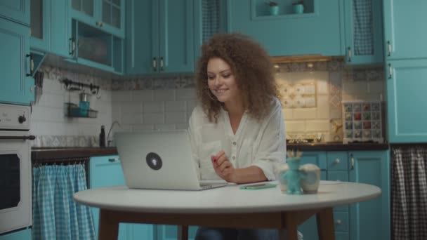 Young curly hair 20s woman finishing her work on laptop and putting hands up with satisfaction, sitting at table on blue kitchen.  - Filmmaterial, Video