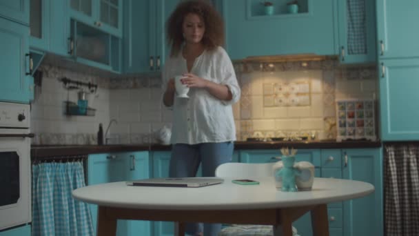 Young curly 20s woman making tea and coming to table with cup. Smiling female opening laptop for working at home on blue kitchen.  - Metraje, vídeo