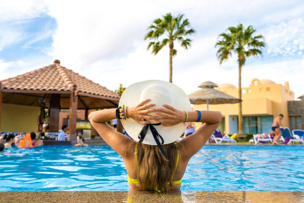 Vue arrière de la jeune femme aux cheveux longs portant un chapeau de paille relaxant en été dans la piscine de l'hôtel avec de l'eau bleue par une journée ensoleillée. - Photo, image