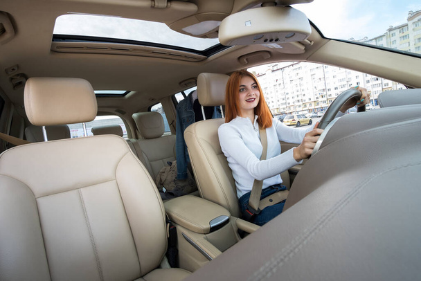 Wide angle view of young redhead woman driver fastened by seatbelt driving a car smiling happily. - Photo, Image