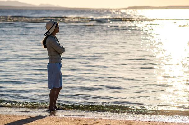 Νεαρή γυναίκα με ψάθινο καπέλο και φόρεμα που στέκεται μόνη της σε άδεια παραλία με άμμο στη θάλασσα. Μοναχικό τουριστικό κορίτσι κοιτάζοντας τον ορίζοντα πάνω από ήρεμη επιφάνεια του ωκεανού για διακοπές ταξίδι. - Φωτογραφία, εικόνα