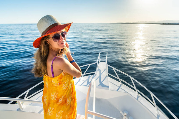 Young woman with long hair wearing yellow dress and straw hat standing on white yacht deck enjoying view of blue sea water. - Zdjęcie, obraz