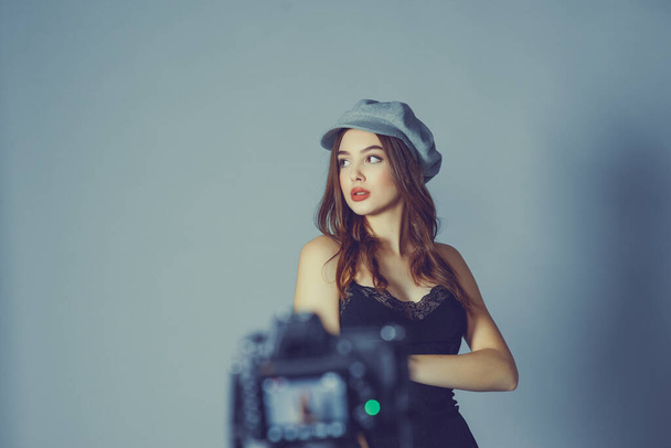Glamorous young woman wears fashionable dress posing with fluttering hair at grey wall.Girl put forward hand in a black dress and a gray cap.A girl is photographed on a camera or I am shooting a video - Photo, Image