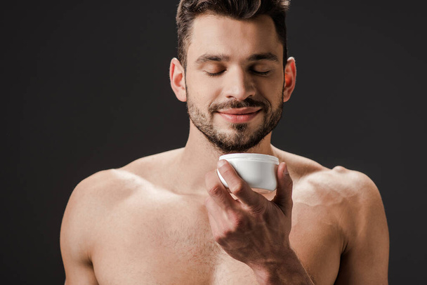 positive naked man with closed eyes holding plastic container with face cream isolated on grey - Photo, Image