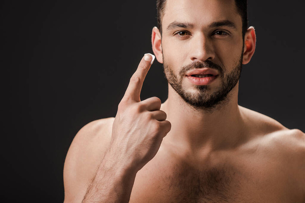 όμορφος γυμνός άντρας απλώνει κρέμα προσώπου που απομονώνεται σε μαύρο - Φωτογραφία, εικόνα