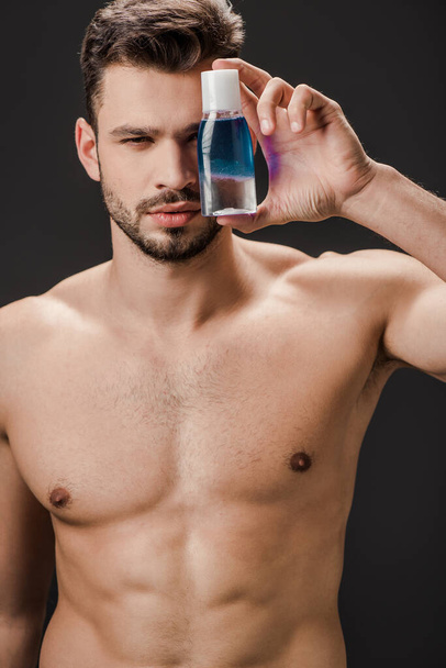 sexy desnudo hombre celebración botella con cosmético líquido aislado en negro
 - Foto, Imagen