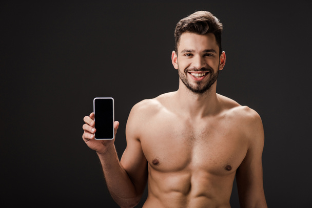 usměvavý sexy nahý muž ukazující smartphone s prázdnou obrazovkou izolované na černé  - Fotografie, Obrázek