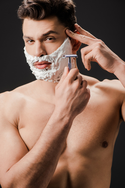 сексуальный обнаженный бородатый мужчина бреющий лицо бритвой изолированный на черном
 - Фото, изображение