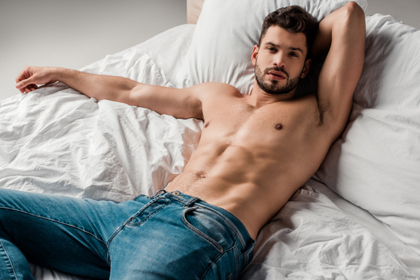 handsome sexy shirtless man in jeans lying on bed on grey - Foto, imagen