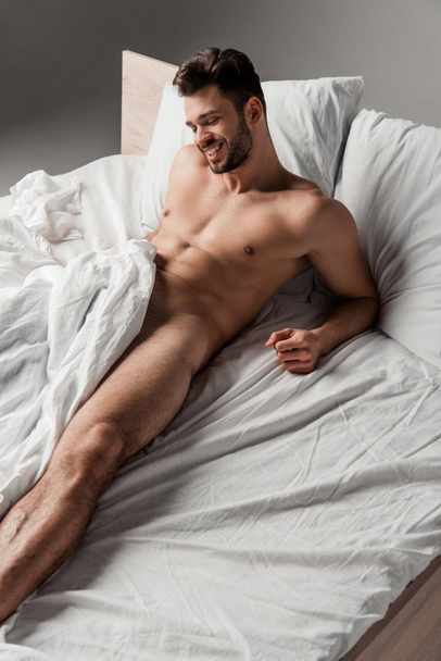 cheerful sexy naked man lying on bed on grey - Photo, Image