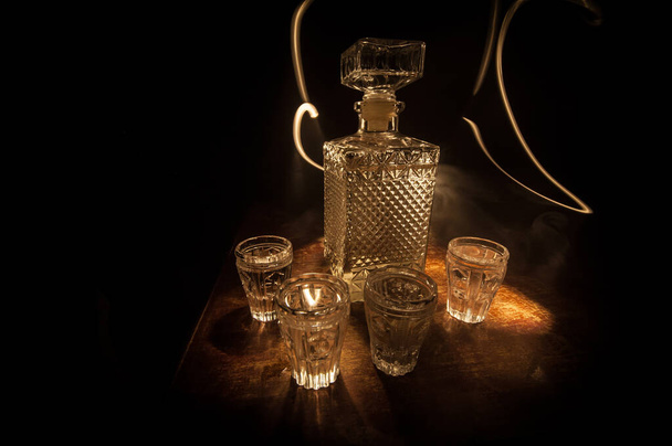 A beautiful carafe with vodka stands on a wooden table with glasses. Dramatic toned foggy background with light. Selective focus. Club drink concept. - Foto, immagini
