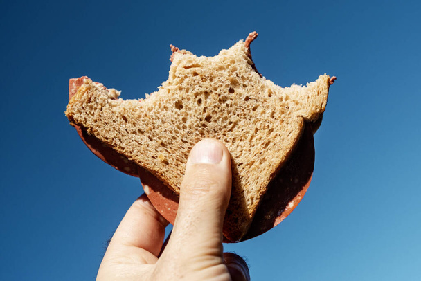 primo piano della mano di un giovane che tiene in mano un panino al salame vegano morso, fatto con pane intero a fette, contro il cielo blu
 - Foto, immagini