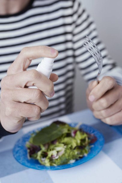 closeup of a caucasian man, sitting at a table set for dinner or lunch, disinfecting a fork by spraying a sanitizer from a white bottle - Photo, Image