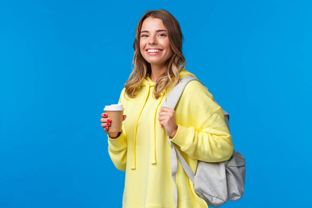 Joyful young blond female student with backpack smiling camera satisfied as holding cup of take-away coffee from favorite cafe after classes, drinking beverage, stand blue background - Photo, Image