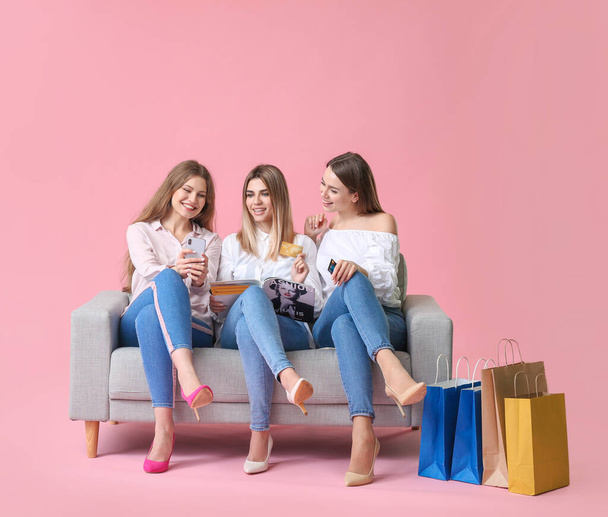 Jovens mulheres com sacos de compras, cartões de crédito e telefone celular sentado no sofá contra fundo de cor
 - Foto, Imagem