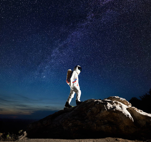 宇宙旅行者の完全な長さは星と息をのむような夜空の下で岩の山に登る。宇宙服を着た宇宙飛行士が巨大な岩の頂上に到達した。天の川銀河と宇宙探査の概念. - 写真・画像