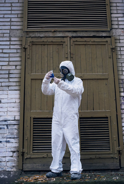Image complète de l'écologiste debout devant une vieille porte et observant la réaction dans une fiole en verre avec un liquide bleu, expérience de laboratoire. Scientifique en uniforme blanc, gants et masque à gaz
 - Photo, image