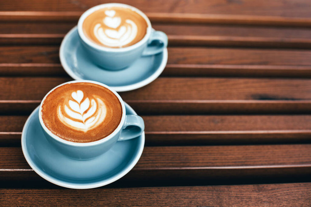 Dva šálky cappuccino s latte art na dřevěném pozadí. Krásná pěna, modré keramické poháry, diagonální složení, místo pro text. - Fotografie, Obrázek