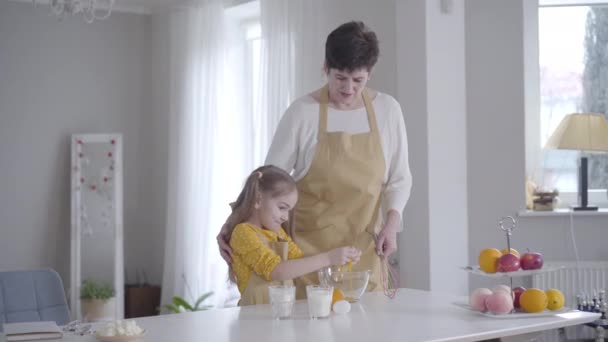 Little cute Caucasian girl breaking egg into bowl and smiling. Happy granddaughter helping grandmother to cook pancakes for Shrove Tuesday. lifestyle, baking, cooking, Shrovetide. - Filmagem, Vídeo