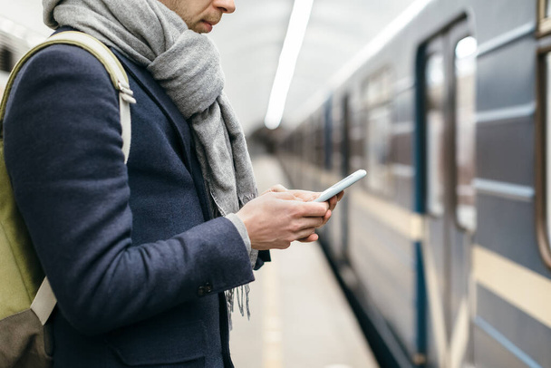 Close up portrait of attractive man in blue coat who looks at his phone and writes a text message. Selective focus. Social networks addiction. Waiting for the train at subway station platform. - 写真・画像