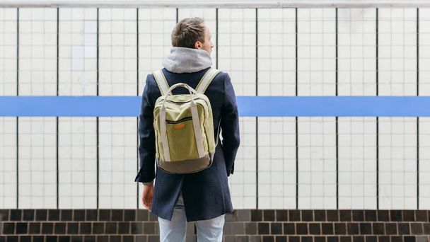 Solitario adulto bell'uomo in un cappotto blu con uno zaino verde dietro in piedi sul marciapiede in attesa del treno. Concentrazione selettiva sul maschio. Ricevuto. Buongiorno, pendolare. Concetto di vita urbana. - Foto, immagini