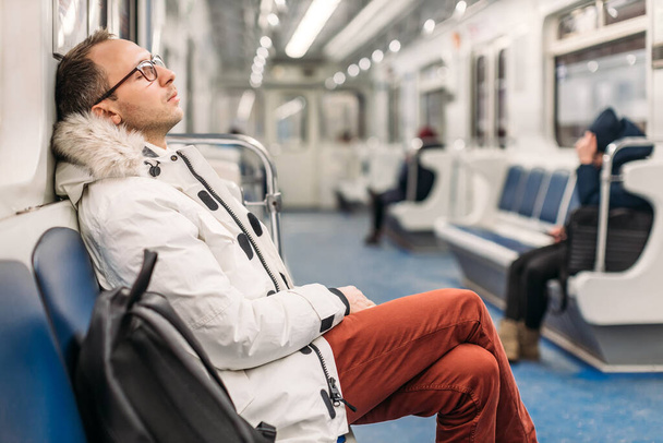 Handsome man with glasses in white jacket and maroon trousers fell asleep on the subway wagon on his way home. Next to the man is a black backpack. Hard working day, stress, fatigue, overwork at work. - Fotó, kép