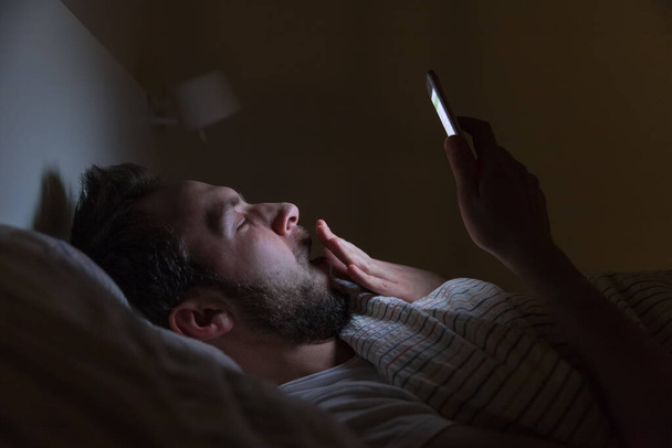 Adult sleepy yawning man awake late at night in bed surfing in web/ sleepy tired, social media addiction, dependency on a cell phone, half-closed tired eyes, negative effect on the eyes, sleeplessness - Photo, Image