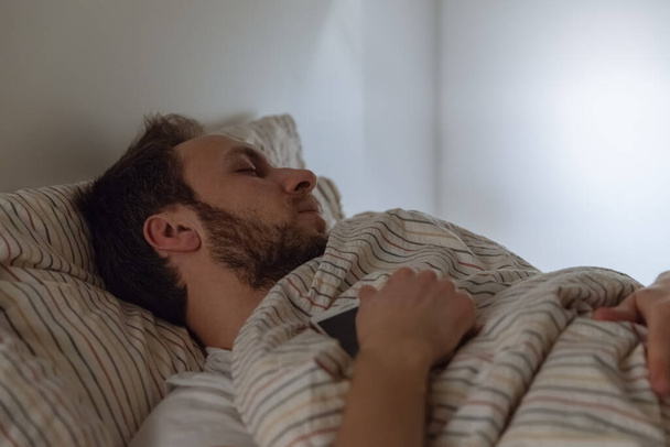 Portret ubranego dorosłego mężczyzny zasnął w łóżku z telefonem w ręku w wyniku zmęczenia / uzależnienia od mediów społecznościowych, wyczerpany mężczyzna, nagromadzone zmęczenie, za mało snu, koncepcja opieki zdrowotnej. - Zdjęcie, obraz