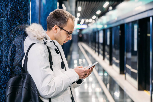 Egy jóképű, fehér kabátos, szemüveges felnőtt férfi fekete hátizsákot hord a vállán. Míg a férfi metrókocsiban várakozik, mobilon üti az időt. Internetes csapda, közösségi média-függőség. - Fotó, kép