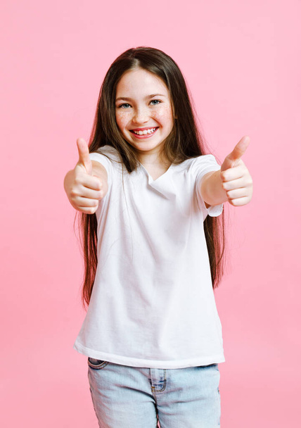 Portrait of adorable smiling little girl child in jeans and white t-shirt with two fingers up isolated on a pink background - Фото, изображение