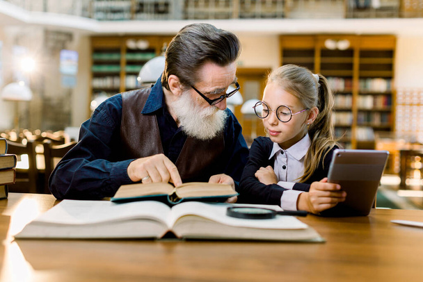 Little pretty girl and her handsome bearded grandfather sitting in vintage library, reading books, using digital tablet. Concept of education for all generations and age - Foto, Imagen
