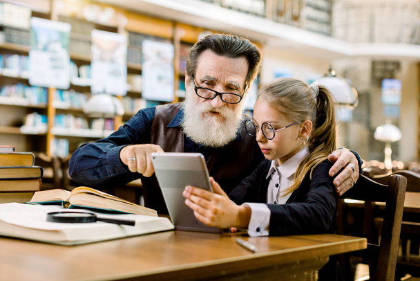 Senior man grandfather and little cute girl granddaughter looking at a tablet, while sitting at the table in old stylish library - Fotoğraf, Görsel