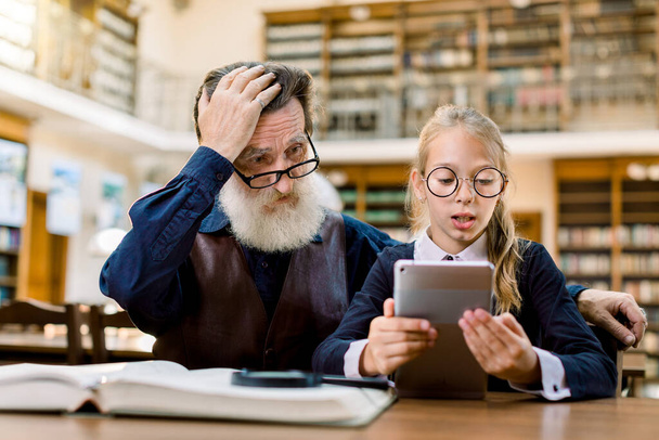 Elegante hombre mayor con su nieta está utilizando una tableta digital en la biblioteca. Chica lee información de la tableta, y el abuelo está confundido y sorprendido
 - Foto, Imagen