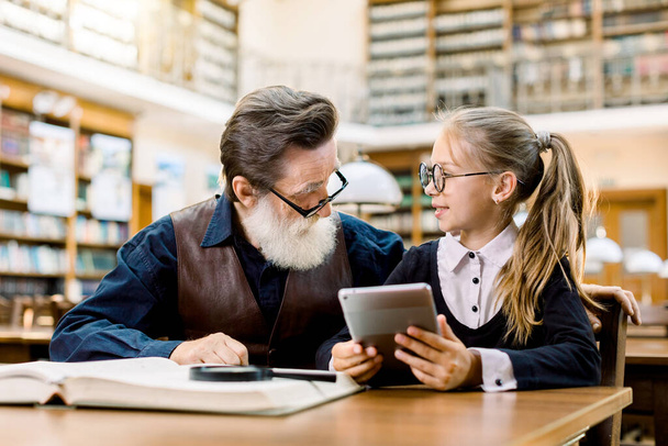 Smart kid girl in eyeglasses sitting at the table in old library and holding tablet, showing something on tablet to her teacher or grandfather. Senior man with his student little girl in library - Photo, image