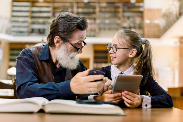 Handsome senior man and little cute girl sitting together in vintage library, compare books, smartphone and new reading digital book device. Grandfather and granddaughter in library - Foto, afbeelding