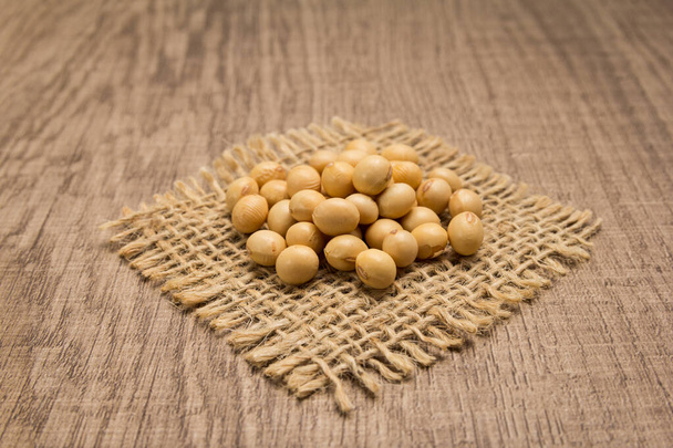 Glycine max ist der wissenschaftliche Name von Sojabohnen-Hülsenfrüchten. Auch bekannt als Sojabohnen und Soja. Körner auf quadratischem Ausschnitt aus Jute. Holztisch vorhanden. Selektiver Fokus. - Foto, Bild