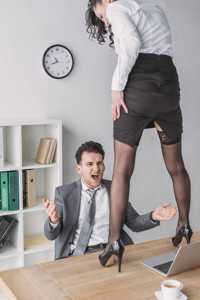 homem de negócios irritado gritando enquanto olha para secretário sexy em pé na mesa em sapatos de salto alto
 - Foto, Imagem