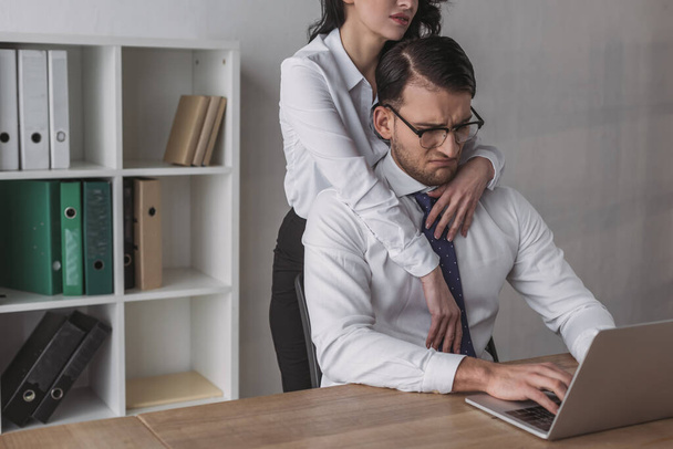 обрезанный взгляд на сексуальную деловую женщину, обнимающую отчаявшегося коллегу, работающего над ноутбуком
 - Фото, изображение