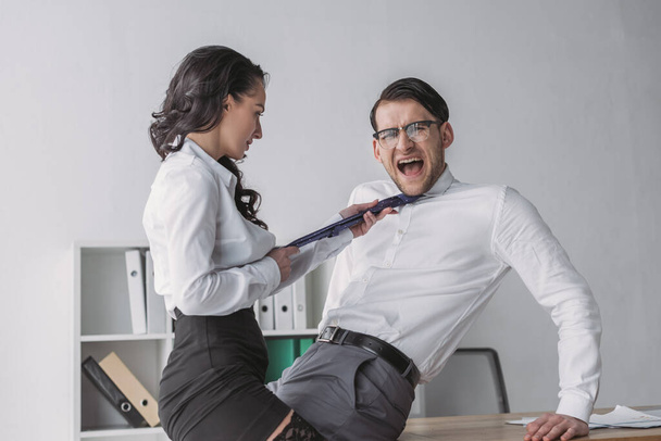 vášnivá podnikatelka dotýkající se kravaty šokovaného kolegy, zatímco svůdný ho v úřadu - Fotografie, Obrázek