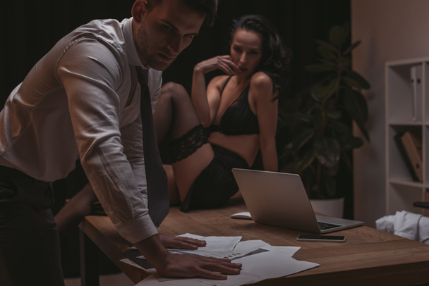 serieus zakenman op zoek naar documenten terwijl sexy secretaresse zitten op bureau in lingerie - Foto, afbeelding