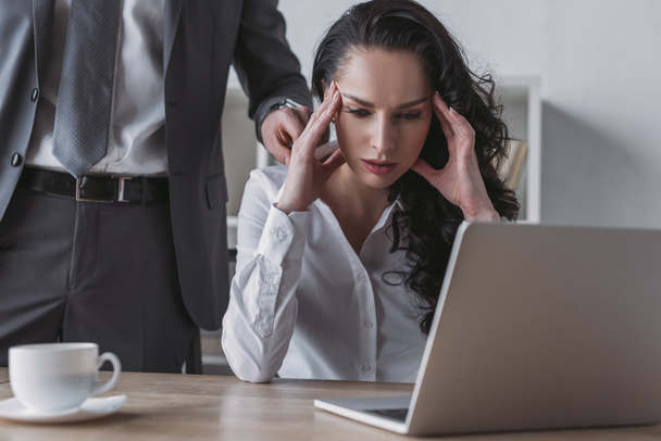 cropped view of businessman touching shoulder of tired secretary sitting at laptop - Photo, Image