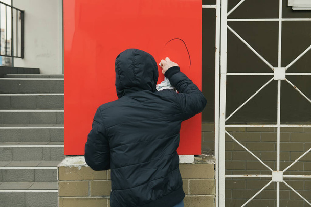 Ismeretlen férfi kék kabátban, csuklyában eltávolítja a vandálok által hagyott foltokat a boltjából. A személy rongyot és mosószert használ. - Fotó, kép