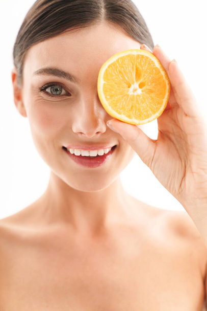 Beauty portrait of an attractive smiling young topless brunette woman standing isolated over white background, showing sliced orange - 写真・画像