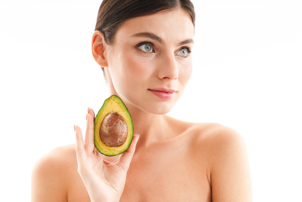 Beauty portrait of an attractive young topless brunette woman standing isolated over white background, showing sliced avocado - Фото, изображение