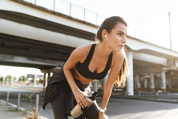 Image of a pretty fitness woman outdoors listening music with earphones drinking water. - Фото, изображение